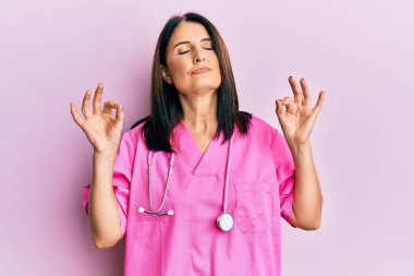 Orta yaşlı esmer kadın doktor üniforması giyiyor ve steteskop takıyor gözleri kapalı gülümsüyor parmaklarıyla meditasyon hareketi yapıyor. yoga konsepti. 
