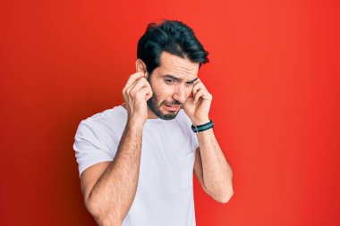 Gündelik beyaz tişört giyen genç İspanyol adam yüksek sesli müzik sesi için kulaklarını parmaklarıyla kapatıyor. sağır kavramı. 