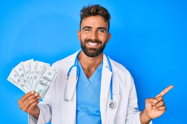 Joven Hispano Vistiendo Uniforme Médico Sosteniendo Dólares Sonriendo Feliz Señalando — Foto de Stock