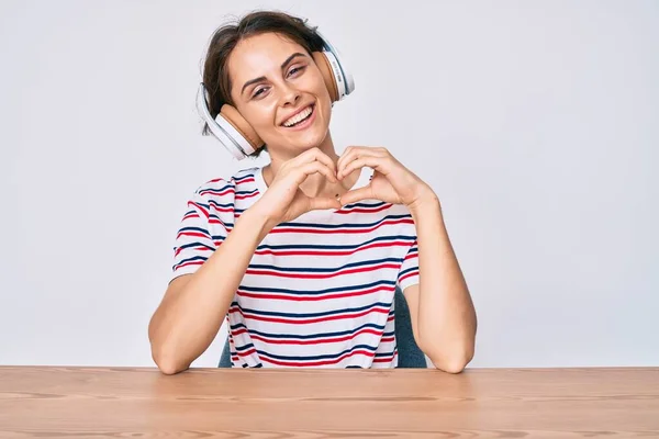 テーブルの上に座っているヘッドフォンを使用して若いヒスパニック系の女性は 手で心のシンボルと形状を示す愛に笑みを浮かべて ロマンチックなコンセプト — ストック写真
