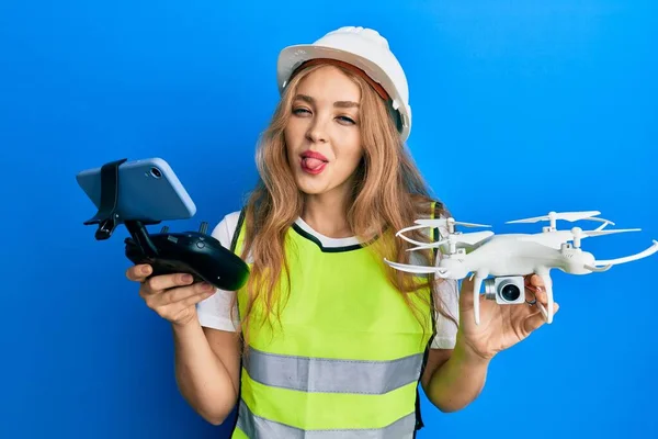 Beautiful blonde caucasian woman wearing safety hard using drone sticking tongue out happy with funny expression.
