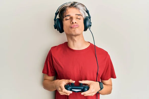 Young hispanic man playing video game holding controller looking at the camera blowing a kiss being lovely and sexy. love expression.