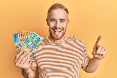 Young caucasian man holding australian dollars smiling with an idea or question pointing finger with happy face, number one 