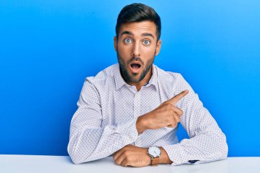 Handsome hispanic man wearing business clothes sitting on the table surprised pointing with finger to the side, open mouth amazed expression. 