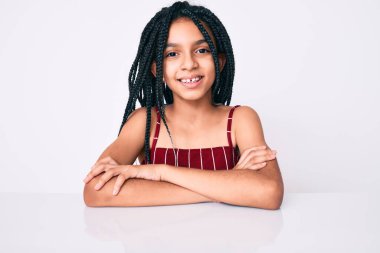 Saçları örgülü, masanın üzerinde oturan, kollarını kavuşturup kameraya bakan mutlu yüzlü Afro-Amerikan kız çocuğu. pozitif kişi. 