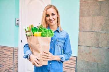 Genç sarışın kız gülümsüyor. Elinde alışveriş poşeti, şehirde duruyor..