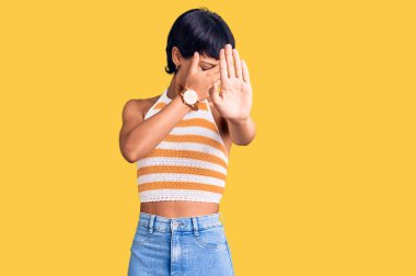 Kısa saçlı, esmer, sıradan kıyafetler giyen, elleriyle gözleri kapayan, üzgün ve korku dolu hareketler yapan genç bir kadın. utangaç ve olumsuz kavram. 