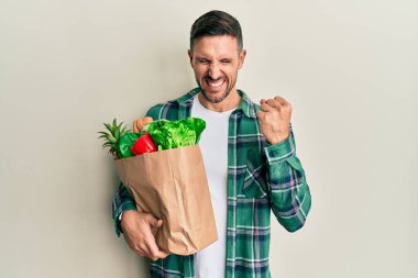 Handsome man with beard holding paper bag with groceries very happy and excited doing winner gesture with arms raised, smiling and screaming for success. celebration concept. 