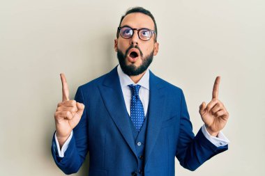 Sakallı, takım elbiseli ve kravatlı genç adam şaşırmış ve şaşırmış bir şekilde yukarı bakıyor ve parmaklarıyla işaret ediyor ve kollarını kaldırıyor.. 