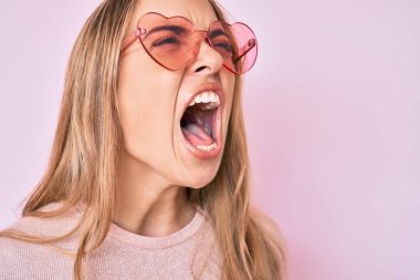 Kalp şeklinde güneş gözlüğü takan genç, sarışın bir kadın kızgın ve kızgın bağırıyor öfkeyle bağırıyor. öfke ve agresif konsept. 