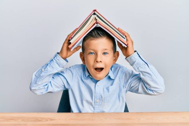 Sevimli beyaz çocuk kafasında kitap tutuyor çılgınlığı kutluyor ve başarıya hayret ediyor gözleri açık heyecanla haykırıyor. 