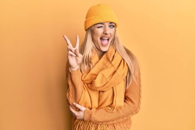 Genç, beyaz kadın, yün kazak ve şapkalı, gülen yüzüyle kameraya bakıp zafer işareti yapan. İki numara.. 