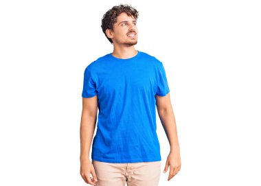 Kıvırcık saçlı, sıradan kıyafetler giyen, yüzünde gülümseme, doğal bir ifade olan genç yakışıklı bir adam. Kendinden emin gülen. 