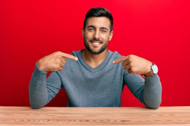 Handsome hispanic man wearing casual style sitting on the table looking confident with smile on face, pointing oneself with fingers proud and happy. 