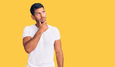 Elleri çenesinde gündelik kıyafetler giyen, soru soran, düşünceli, genç Latin bir adam. Düşünceli bir yüzle gülümsüyor. şüphe kavramı. 