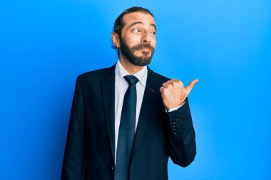 Uzun saçlı, sakallı, takım elbiseli, kravatlı, gülen yüzlü ve baş parmağıyla yüzünü gösteren çekici bir adam.. 