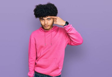 Young african american man with afro hair wearing casual pink sweatshirt pointing unhappy to pimple on forehead, ugly infection of blackhead. acne and skin problem 
