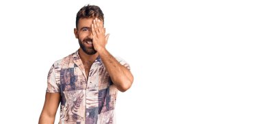 Young hispanic man wearing summer clothes covering one eye with hand, confident smile on face and surprise emotion. 