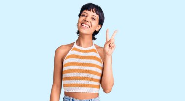 Kısa saçlı, sıradan kıyafetler giyen, gülümseyen, parmaklarını zafer işareti yaparken gösteren genç esmer kadın. İki numara.. 