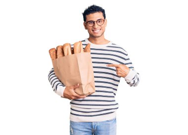 İspanyol asıllı yakışıklı genç adam, elinde kağıt torba, elinde ekmek, gülümsüyor, mutlu bir şekilde elini ve parmağını gösteriyor. 