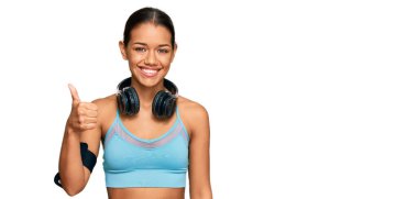 Beautiful hispanic woman wearing sportswear and headphones smiling happy and positive, thumb up doing excellent and approval sign 