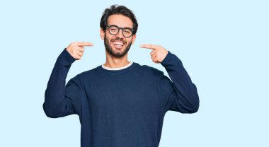 Gündelik kıyafetler giyen ve gözlüklü, neşeli gülümseyen, parmaklarıyla ve ağzıyla işaret eden İspanyol genç bir adam. diş sağlığı kavramı. 