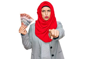 Beautiful hispanic woman wearing islamic hijab holding australian dollars banknotes pointing with finger to the camera and to you, confident gesture looking serious 