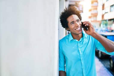 Gülümseyen mutlu gülümseyen Afrikalı Amerikalı genç bir adam. Yüzünde gülümsemeyle duvara yaslanarak şehrin caddesinde akıllı telefondan konuşarak..