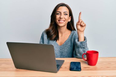 Ofiste çalışan esmer bir kadın bilgisayarlı dizüstü bilgisayar kullanıyor. Bir fikir veya soruyla gülümsüyor ve mutlu bir yüzle işaret ediyor. 