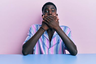 Gündelik giysiler giymiş genç Afro-Amerikan bir adam masada oturuyor ağzı yanlışlıkla ellerle kapatırken şok geçiriyor. gizli kavram. 