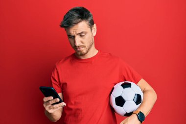 Elinde futbol topu tutan yakışıklı beyaz adam akıllı telefondan kuşkucu ve gergin bakıyor, sorun yüzünden surat asıyor. negatif kişi. 