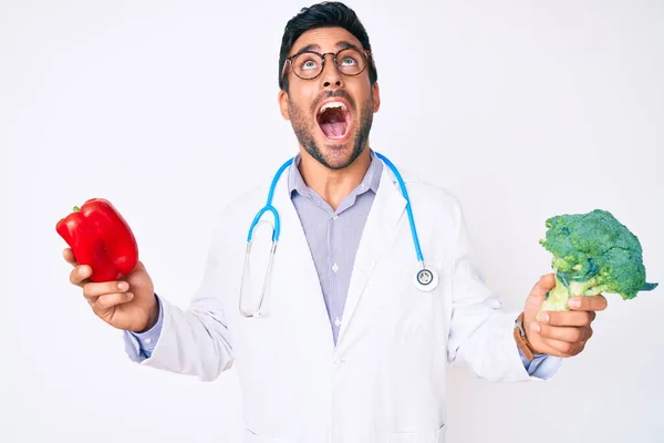 Young Hispanic Man Wearing Doctor Stethoscope Holding Red Pepper Broccoli — Fotografia de Stock