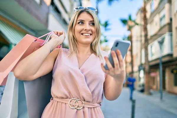Młoda Blondynka Uśmiecha Się Szczęśliwy Trzymając Torby Zakupy Pomocą Smartfona — Zdjęcie stockowe