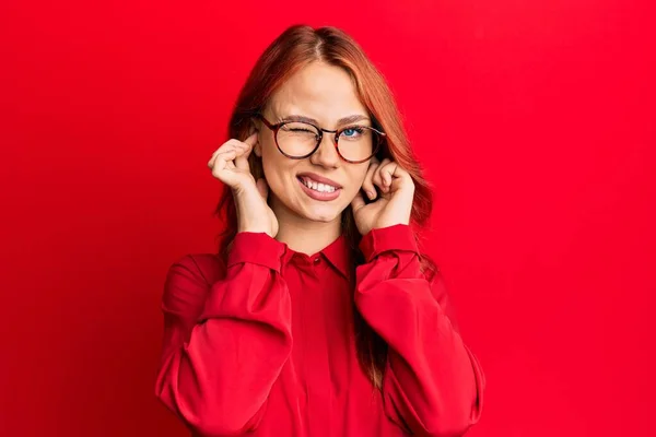 赤い背景にカジュアルな服と眼鏡を身に着けている若い美しい赤毛の女性は 大きな音楽のノイズのためにいらいらした表情で指で耳をカバーしています 聴覚障害の概念 — ストック写真