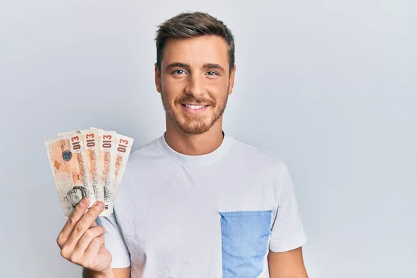 Handsome caucasian man holding 10 united kingdom pounds banknotes looking positive and happy standing and smiling with a confident smile showing teeth