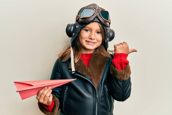 Little beautiful girl wearing pilot uniform holding paper plane pointing thumb up to the side smiling happy with open mouth 