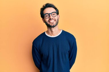 Gündelik kıyafetler ve gözlük takan genç İspanyol adam pozitif ve mutlu görünüyor ve kendine güvenen bir gülümsemeyle dişlerini gösteriyor. 