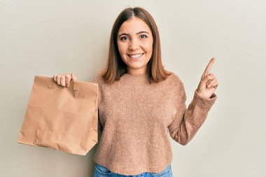 Elinde kağıt poşet tutan genç ve güzel bir kadın bir fikirle ya da soruyla gülümsüyor ve mutlu bir yüzle işaret ediyor. 