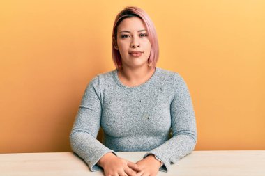 Pembe saçlı, sıradan kıyafetler giyen İspanyol kadın masada oturuyor ve yüzünde ciddi bir ifade var. Basit ve doğal bir şekilde kameraya bakıyorsun.. 