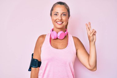 Orta yaşlı İspanyol kadın spor kıyafetleri giyiyor ve kulaklık takıyor. Kameraya gülümsüyor ve zafer işareti yapıyor. İki numara.. 