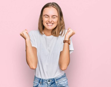 Başarı için heyecanlı, kollarını kaldırmış ve gözleri kapalı, gündelik beyaz tişört giyen güzel sarışın kadın zaferi kutluyor. Kazanan konsept. 