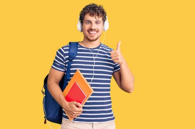 Kıvırcık saçlı, öğrenci çantası ve kitaplarıyla gülümseyen, mutlu ve olumlu gülümseyen, başparmağıyla mükemmel ve onay işareti yapan genç ve yakışıklı bir adam. 