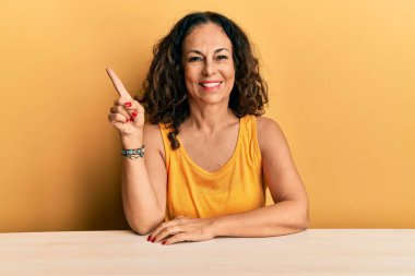 Masada oturan, yüzünde kocaman bir gülümseme olan orta yaşlı, güzel bir kadın. Elinle kameraya bakıyor.. 