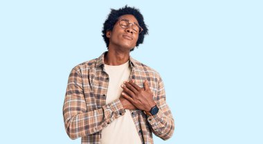 Afro saçlı, sıradan giyinmiş, gözlüklü, göğsünde elleri kapalı, yüzünde minnet ifadesi olan yakışıklı Afro-Amerikan erkeği. sağlık kavramı. 