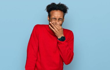 Young african american man with beard wearing casual winter sweater smelling something stinky and disgusting, intolerable smell, holding breath with fingers on nose. bad smell 