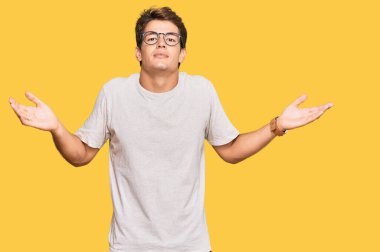 Sıradan kıyafetler giyen, gözlüklü, kafası karışık, elleri ve kolları havada yakışıklı beyaz bir adam. şüphe kavramı. 