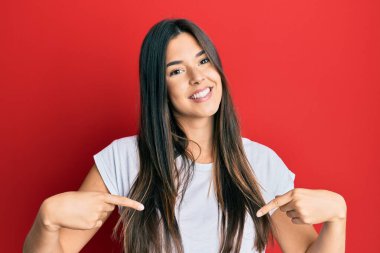 Genç esmer kadın kırmızı arka planda günlük beyaz tişört giyiyor. Kendine güvenen, yüzünde gülümseme olan, kendini gururlu ve mutlu gösteren bir kadın.. 