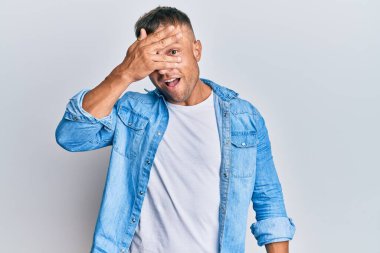 Handsome muscle man wearing casual denim jacket peeking in shock covering face and eyes with hand, looking through fingers with embarrassed expression. 