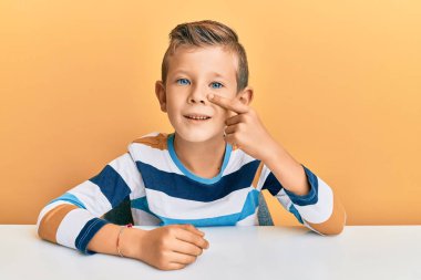 Kafkasyalı sevimli bir çocuk, günlük giysiler giymiş, masaya oturmuş, parmaklarıyla yüzünü ve burnunu göstererek, neşeli bir şekilde gülümsüyor. güzellik konsepti 