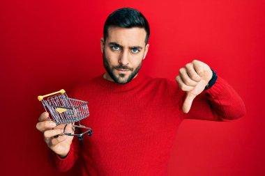 Genç İspanyol adam elinde kızgın suratlı küçük bir süpermarket arabası tutuyor. Negatif işareti baş parmakları aşağıda, reddedilme konsepti ise... 